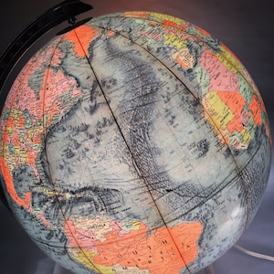 Może przedstawiać: Antyczny globus z czarną metalową podstawą. Globus jest podświetlony od wewnątrz, co sprawia, że kontynenty i oceany świecą w odcieniach niebieskiego, zielonego i żółtego. Globus zawiera szczegółową mapę z nazwami krajów i granicami.