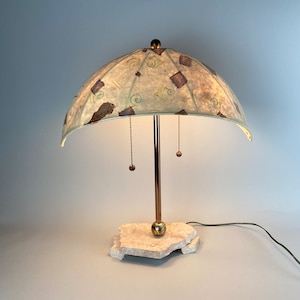May include: A table lamp with a white and green patterned lampshade. The lampshade is shaped like an umbrella and has a gold metal base. The lamp is sitting on a white stone base.