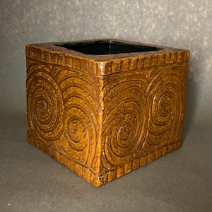 May include: A square, brown ceramic planter with a textured surface. The planter features a repeating spiral design carved into the sides. The interior is a dark, glossy color. The planter is a decorative item.