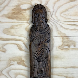 May include: A dark brown wooden carving of a person with a beard and long hair, wearing a robe. The carving is detailed and intricate, with a realistic expression on the person's face.