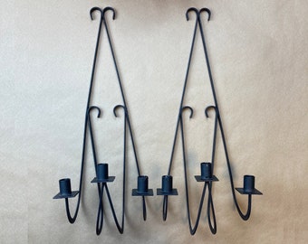 May include: A set of three black metal candle holders with a decorative design. Each holder has two arms that curve upwards and outwards, creating a unique shape. The holders are designed to hold candles in the center of the arms.