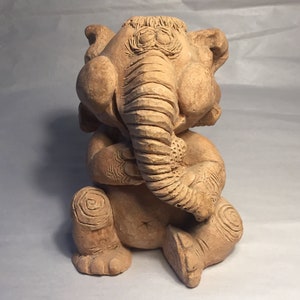 May include: A terracotta sculpture of an elephant sitting with its trunk curled up in front of its face. The elephant has a detailed, textured surface and a whimsical expression.
