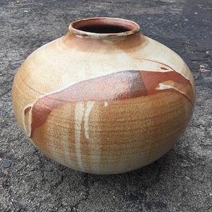 Puede incluir: Un jarrón grande, redondo y de cerámica con un esmalte marrón, blanco y naranja. El jarrón tiene una boca ancha y un fondo redondeado.