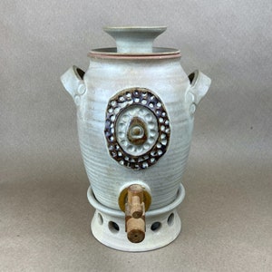 May include: A white ceramic pitcher with a brown and white design. The pitcher has a wooden spigot and a lid.