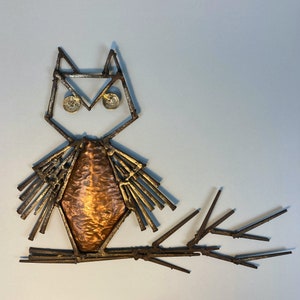 May include: A metal owl sculpture with a copper-colored body and a wire frame. The owl has large, round eyes and a long, thin beak. The sculpture is mounted on a wire base.