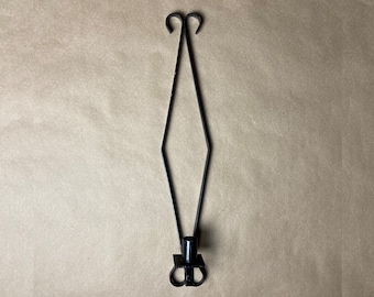 May include: Black metal candle holder with a diamond-shaped design. The holder has two loops at the top and a single candle holder at the bottom.