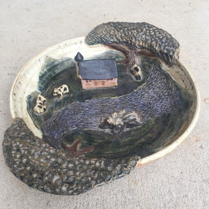 May include: A ceramic decorative plate featuring a rural landscape scene. The plate depicts a small house, a tree, and a river, all in a textured, earthy color palette. The rim of the plate has a textured, stone-like appearance.