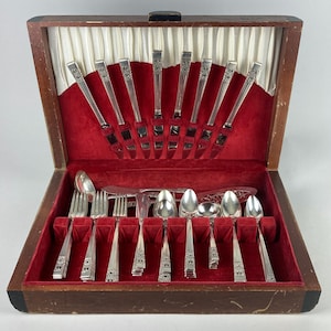 May include: A vintage silverware set in a wooden case with a red velvet interior. The set includes knives, forks, spoons, and serving utensils. The silverware has a decorative pattern on the handles and is arranged neatly within the case.