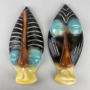May include: Two ceramic wall masks with a tribal design. Each mask has a black and brown top, blue eyes, a brown nose, and a yellow base. Silver accents highlight the details of the masks.