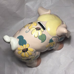 Puede incluir: Una alcanc&iacute;a de cer&aacute;mica rosa con un dise&ntilde;o floral amarillo y verde. La alcanc&iacute;a tiene una ranura en la parte posterior para monedas.
