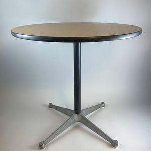 Op de afbeelding: Een ronde tafel van licht hout met een zwarte rand en een zwarte metalen voet. De voet heeft een vijfpuntige stervorm met zilverkleurig metaal.