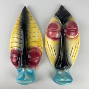 May include: Two ceramic tribal masks with a leaf-like shape. The masks feature a color palette of yellow, black, red, and blue. The masks have a stylized face with closed eyes and a long nose. The masks are likely for wall decor.
