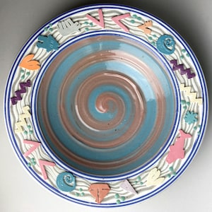 May include: A decorative ceramic bowl with a spiral design in shades of blue and brown. The rim features colorful geometric shapes and a wavy pattern. The bowl has a blue and white striped border.
