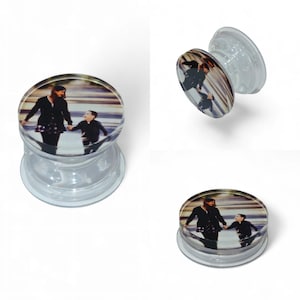 May include: Clear plastic phone grip with a circular image of a person and a child holding hands. The grip has a collapsible design for easy use and a flat base for secure placement on a phone.