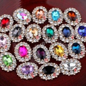 Op de afbeelding: Een verzameling ovale edelstenen in verschillende kleuren, waaronder rood, roze en blauw, elk omringd door kleine, heldere kristallen. De edelstenen zijn gezet in een goudkleurige metalen basis.