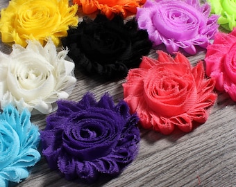 May include: A collection of colorful fabric flower embellishments in various shades, including white, yellow, orange, black, purple, red, pink, and blue. The flowers have a ruffled petal design and are arranged on a wooden surface.