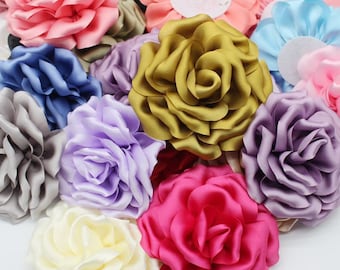 May include: Assortment of fabric flower embellishments in various colors, including pink, purple, blue, gold, cream, and gray. The flowers are made of satin and have a layered, ruffled appearance.