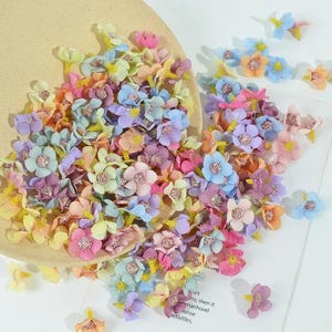 May include: A collection of small, pastel-colored artificial flowers in shades of pink, blue, yellow, and purple. The flowers are scattered on a light brown surface and a white surface. Some flowers have glittery centers.