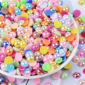 Puede incluir: Primer plano de una colección de cuentas coloridas, semiredondas, con aspecto de perlas. Las cuentas son de varios tonos de rosa, amarillo, naranja, azul y verde. Algunas cuentas tienen un brillo nacarado. Las cuentas están esparcidas en un cuenco blanco.