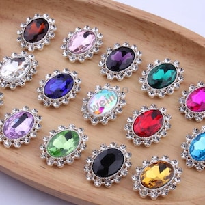 May include: Assortment of oval-shaped rhinestone embellishments in various colours, including clear, pink, purple, green, red, black, and gold. Each embellishment features a silver-toned metal setting with a decorative border of smaller rhinestones.