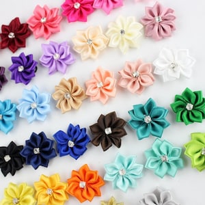 Puede incluir: Surtido de flores de cinta de satén de colores con un centro de strass. Las flores están dispuestas en un patrón aleatorio sobre un fondo blanco.