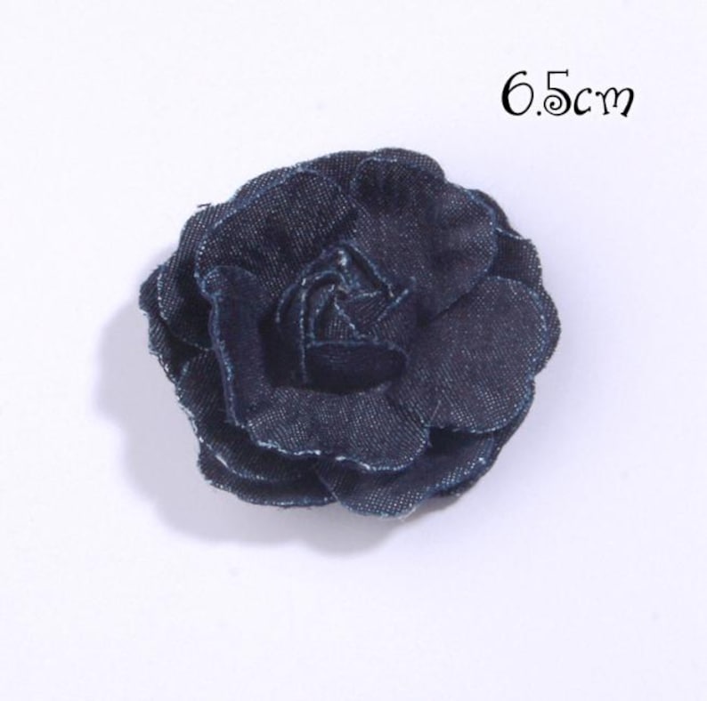 Puede incluir: Una flor de tela vaquera azul oscuro, de 6,5 cm de di&aacute;metro, que se asemeja a una rosa. La flor tiene m&uacute;ltiples p&eacute;talos en capas y un aspecto texturizado. La flor est&aacute; sobre un fondo blanco.