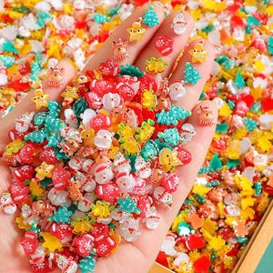 Puede incluir: Una mano sosteniendo una colección de adornos coloridos con temática navideña. El surtido incluye miniaturas de Papá Noel, muñecos de nieve, árboles de Navidad, estrellas y renos en rojo, blanco, amarillo y turquesa. El fondo está lleno de decoraciones similares.