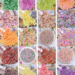 Könnte beinhalten: Eine Sammlung kleiner, farbenfroher Polymer-Ton-Scheiben in verschiedenen Formen und Farben, darunter Früchte und Blumen. Die Scheiben werden in transparenten Plastikgläsern und einer weißen Schüssel präsentiert, ideal für Nagelkunst oder Bastelprojekte.