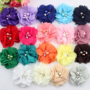 Puede incluir: Una colección de adornos florales de tela en varios tonos, incluyendo morado, rojo, rosa, verde y naranja. Cada flor presenta un grupo de perlas de imitación y pedrería en el centro, añadiendo un toque de brillo.
