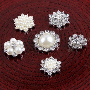 May include: Six silver-toned embellishments with pearl and rhinestone accents. The embellishments are arranged on a red surface.