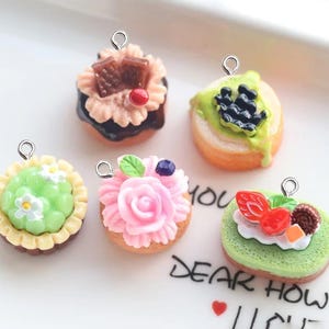 Puede incluir: Cinco encantos de pastel en miniatura con anillos de salto plateados. Los pasteles están decorados con varios ingredientes, que incluyen chocolate, fruta y flores.