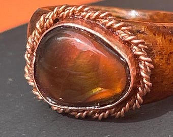 May include: A ring featuring a polished dark brown wooden band and a central teardrop-shaped iridescent gemstone. The stone is encased in a twisted copper wire setting that creates a rope-like border around the perimeter of the gem.