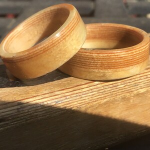 May include: Two wooden rings with a light brown and reddish-brown striped design. The rings are made of layered wood, with the grain visible. The rings are sitting on a wooden surface, and the lighting suggests a sunny day.