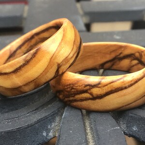 Puede incluir: Dos anillos de madera con un patr&oacute;n de veta de madera natural. Los anillos est&aacute;n hechos de madera de olivo y tienen un color marr&oacute;n claro con vetas marrones m&aacute;s oscuras.