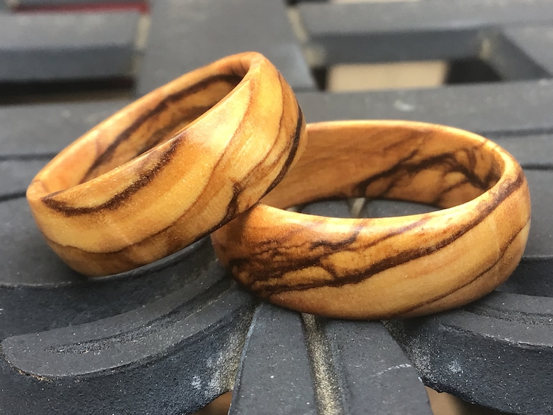 Puede incluir: Dos anillos de madera con un patr&oacute;n de veta de madera natural. Los anillos est&aacute;n hechos de madera de olivo y tienen un color marr&oacute;n claro con vetas marrones m&aacute;s oscuras.