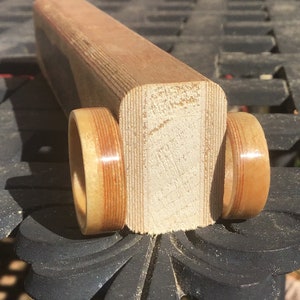 May include: A wooden toy car with two wheels and a rectangular body. The wheels and body are made of light brown wood with darker brown stripes. The toy is sitting on a dark surface.