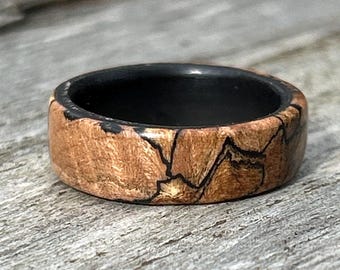 May include: A ring featuring a smooth, polished outer band made of light brown wood with dark, irregular, vein-like patterns running throughout the surface. The interior of the ring is lined with a solid, matte black material.