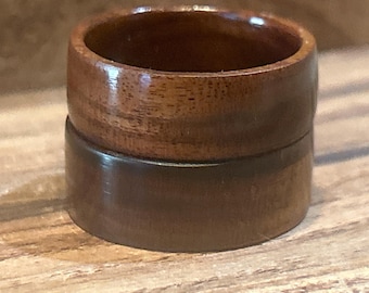 May include: Two stacked rings crafted from polished wood. The top ring features a lighter, reddish-brown wood grain with visible natural patterns, while the bottom ring is composed of a darker, solid brown wood with a smooth, uniform finish.