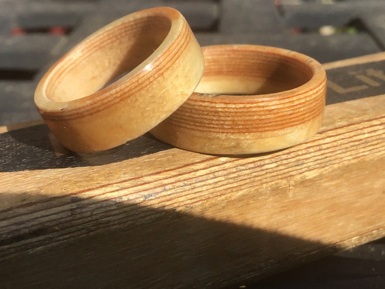 May include: Two wooden rings with a light brown and reddish-brown striped design. The rings are made of layered wood, with the grain visible. The rings are sitting on a wooden surface, and the lighting suggests a sunny day.