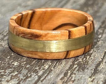 May include: A ring crafted from light brown wood with a prominent, smooth, horizontal band of brushed gold-colored metal running through the center. The wood grain features dark brown streaks and a polished finish, creating a contrast with the metal.