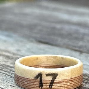 May include: A wooden ring with the number 17 painted in black. The ring has a light-colored top and a darker, layered band around the middle. The ring is set against a blurred wooden surface.