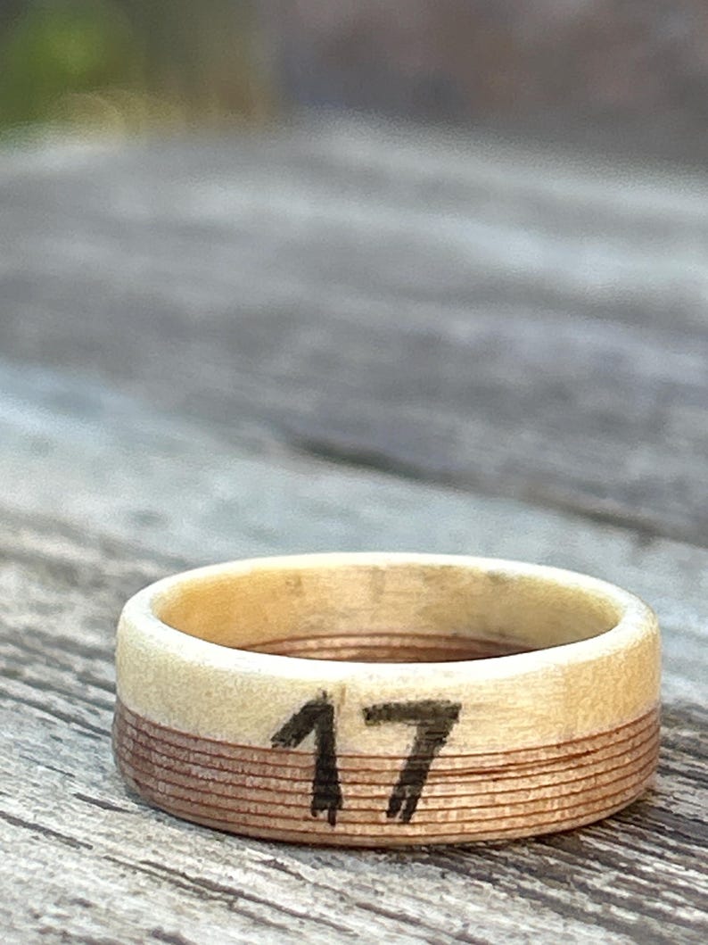 May include: A wooden ring with the number 17 painted in black. The ring has a light-colored top and a darker, layered band around the middle. The ring is set against a blurred wooden surface.