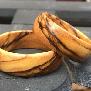 Puede incluir: Dos anillos de madera con un patr&oacute;n de veta de madera natural. Los anillos est&aacute;n hechos de madera de olivo y tienen un color marr&oacute;n claro con vetas marr&oacute;n oscuro.