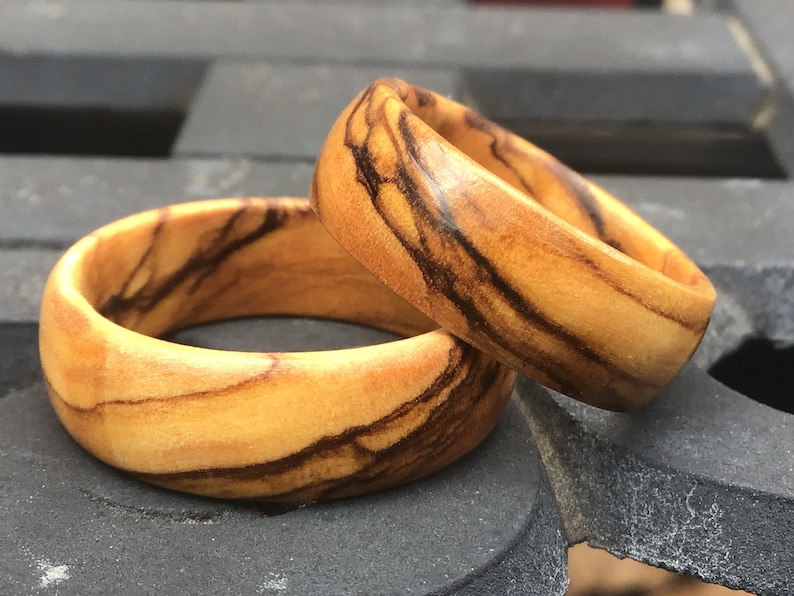 Puede incluir: Dos anillos de madera con un patr&oacute;n de veta de madera natural. Los anillos est&aacute;n hechos de madera de olivo y tienen un color marr&oacute;n claro con vetas marr&oacute;n oscuro.