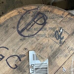 K&ouml;nnte beinhalten: Nahaufnahme eines Holzfasses mit schwarzen Markierungen. Das Fass hat ein Etikett mit dem Text "D431" und einem Barcode. Der Text "Jim Motow, Prop.  Exchequer, Tex." ist auf dem Fass sichtbar.