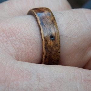 K&ouml;nnte beinhalten: Nahaufnahme eines Holzrings mit einer dunkelbraunen Beize. Der Ring wird an einem Finger getragen.