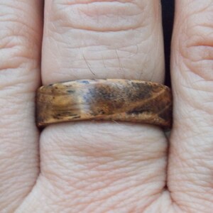 K&ouml;nnte beinhalten: Nahaufnahme eines Holzrings mit braunem und schwarzem Holzmaserungsmuster. Der Ring wird an einem Finger getragen.