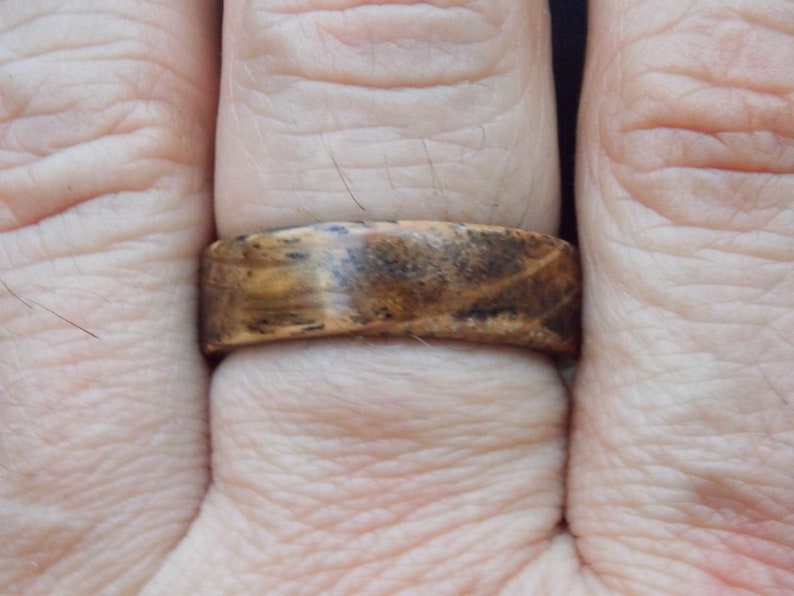 K&ouml;nnte beinhalten: Nahaufnahme eines Holzrings mit braunem und schwarzem Holzmaserungsmuster. Der Ring wird an einem Finger getragen.