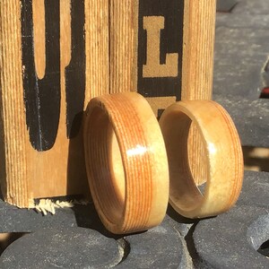 May include: Two wooden rings with a light brown and reddish-brown striped pattern. The rings are displayed on a dark surface with a wooden block in the background. The block has black lettering.