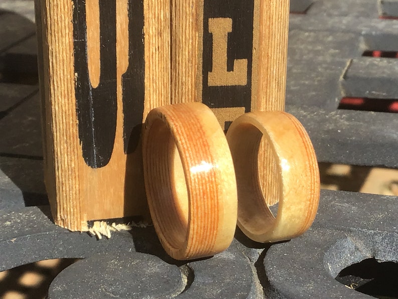 May include: Two wooden rings with a light brown and reddish-brown striped pattern. The rings are displayed on a dark surface with a wooden block in the background. The block has black lettering.